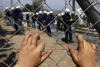A child places his hands on a fence as Greek police officers stand guard at a makeshift camp for migrants and refugees in Greece.